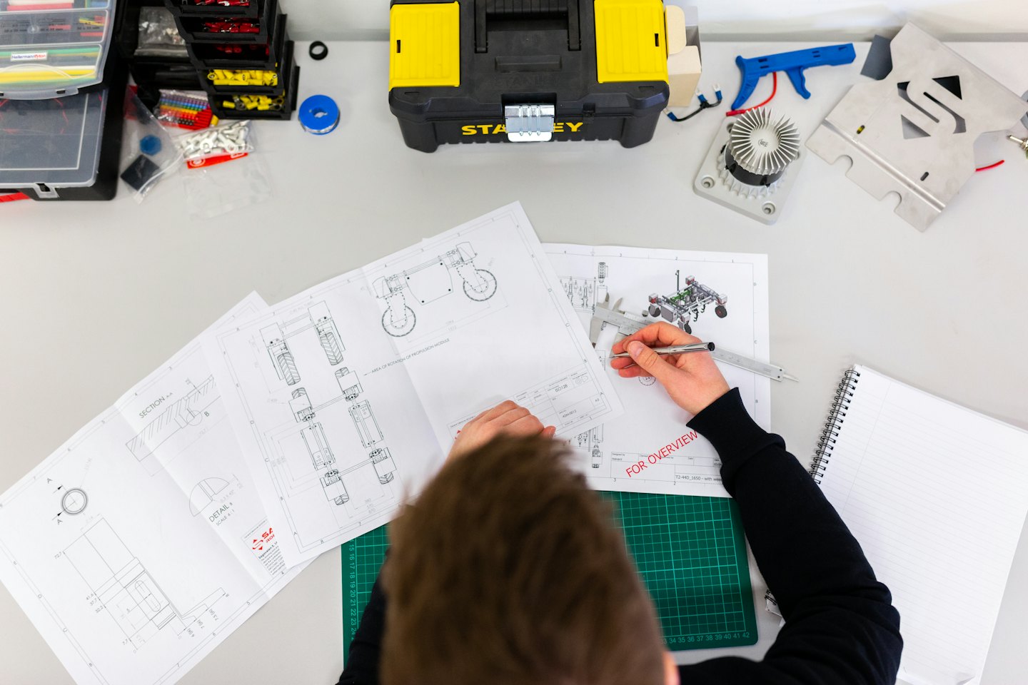 Portfolio Engineer reviews schematics with calipers at a cluttered desk.