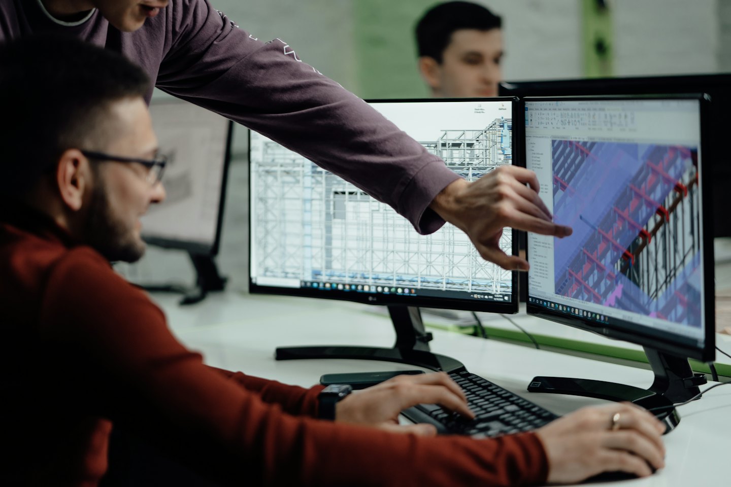 Services Man points at 3D design on dual monitors while colleague types.