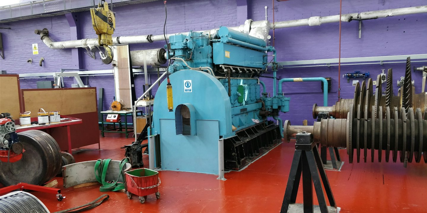 Booking A large industrial engine sits in a workshop on red flooring.